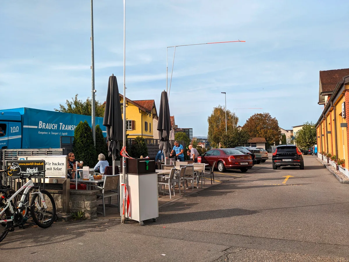 Bisher gab es einige Tische und Stühle neben dem Parkplatz. Geplant ist ein Anbau mit Terrasse und einem kleinen Park. Man sieht den Parkplatz einer Bäckerei.